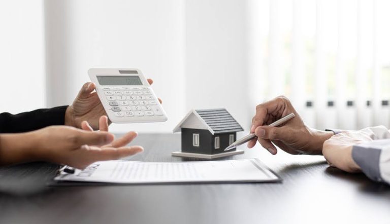 close-up of the hands of two people on opposite sides of a table looking at a calculator and notepad with a model home in the background