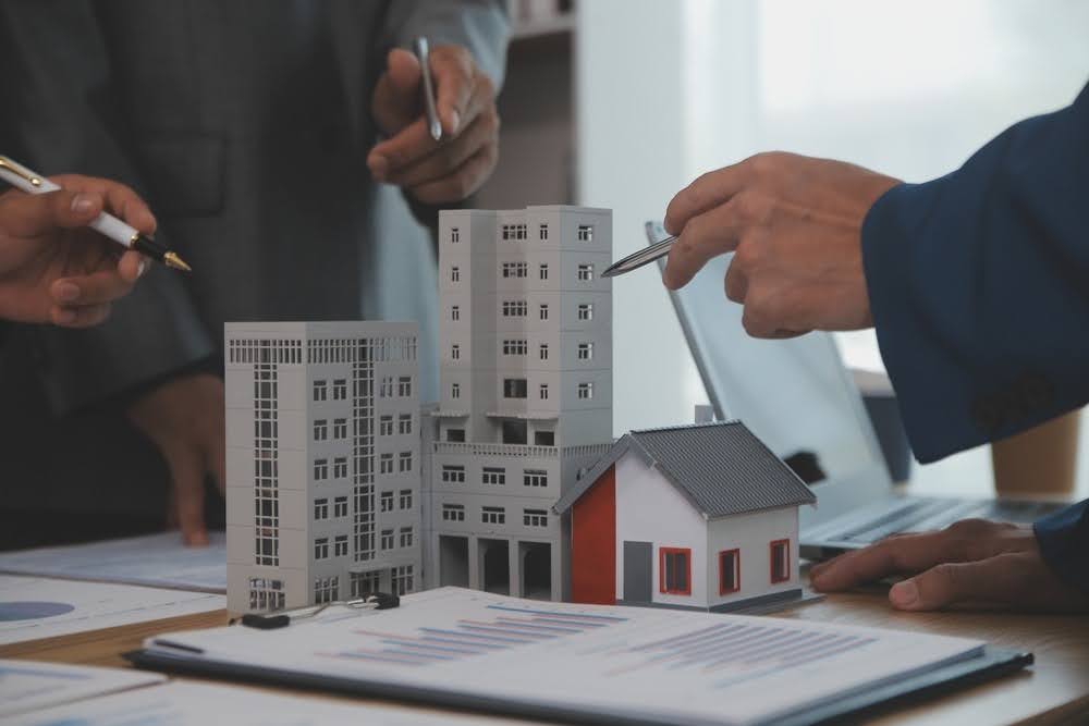 close-up of hands of three men in suits looking at a development model with notebooks and a laptop alongside on the desk
